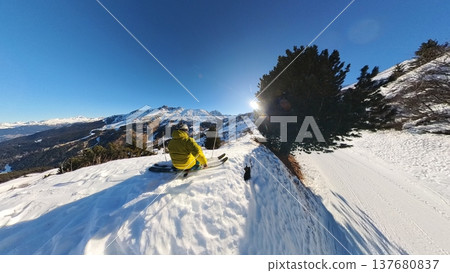 Skier in Yellow Jacket Sitting on Snowy Alpine Ridge Overlooking Valley 137680837