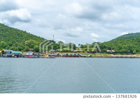 Scenic view of colorful floating wooden rafts and resorts on a peaceful lake surrounded by lush green mountains under a cloudy sky in Thailand. Tropical vacation and travel destination concept. 137681926