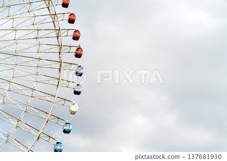 Close-up view of a large Ferris wheel with colorful gondolas against a cloudy sky. Features red, purple, white, and blue cabins on a white metal frame. Minimalist composition with copy space. 137681930