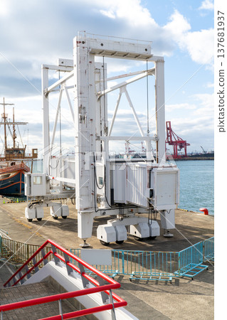 Large white mobile passenger gangway on wheels at a busy harbor. Professional boarding bridge for ships located on a dock under a bright sky with industrial port equipment in the background. 137681937