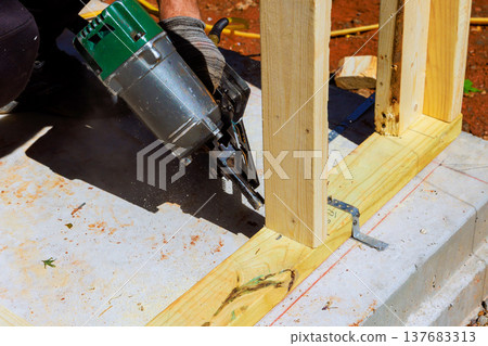 Construction worker uses nail gun to fasten wooden frame on building site Construction worker uses nail gun to fasten wooden frame on building site 137683313