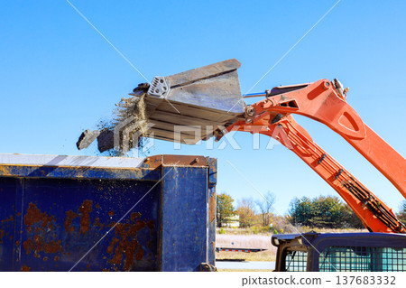 Construction vehicle unloads material into blue dumpster under clear sky at worksite 137683332