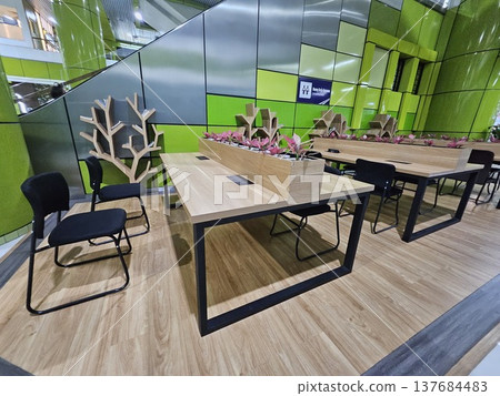 A modern co-working space at a station featuring wooden desks with black chairs, unique tree-shaped bookshelves, and small plants set against a vibrant green wall with integrated power outlets. 137684483