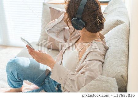 A relaxed woman sitting on a sofa at home, listening to music with headphones on. 137685830