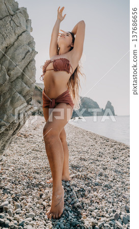 Bikini woman beach posing gracefully beside a large rock on a pebble shore under the bright sun 137686656