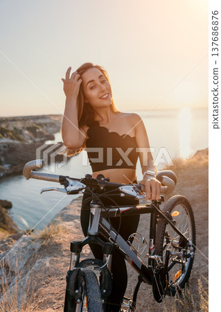 Woman bicycle sunset Young woman posing with her mountain bike on a scenic cliff edge overlooking the sea during golden hour sunset for healthy lifestyle 137686876