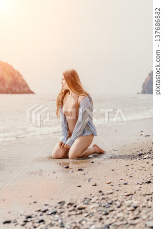 Woman beach sunrise young redhead woman kneeling on sandy shore looking away at the ocean with soft warm light Woman beach sunrise young redhead woman kneeling on sandy shore looking away at the ocean with soft warm light 137686882