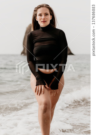 Woman beach swimwear posing in a black mesh top on the shoreline with waves and a rock formation in the background Woman beach swimwear posing in a black mesh top on the shoreline with waves and a rock formation in the background 137686883