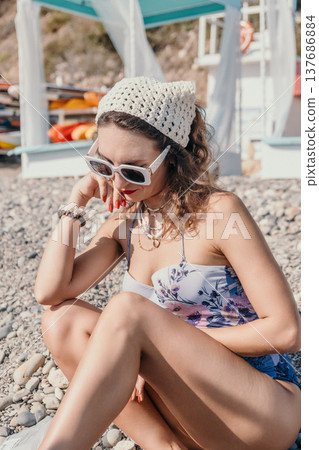 Woman beach summer stylish young lady wearing floral swimsuit and crochet headscarf sitting on pebble shore at sunny resort 137686884