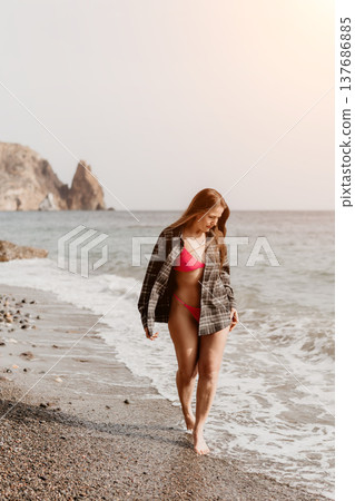 Woman beach vacation young woman walks barefoot on a pebble beach wearing a pink bikini and flannel shirt under the summer sun Woman beach vacation young woman walks barefoot on a pebble beach wearing a pink bikini and flannel shirt under the summer sun 137686885