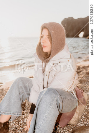 Woman beach sunset sits peacefully on a pebble shore wearing a warm puffer jacket and knitted hood during golden hour 137686886