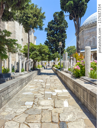 Stone pathway lined with trees and flowers leading to a historic building under a clear blue sky in a serene outdoor setting 137687354