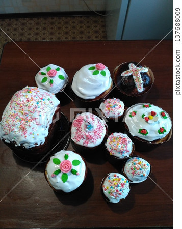 Assorted baked goods with colorful icing and decorations arranged on a wooden table, featuring cakes and cupcakes with floral designs and sprinkles 137688009