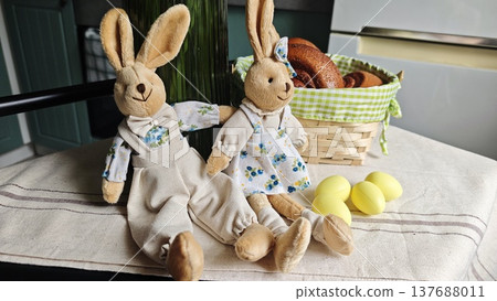 Plush bunny dolls dressed in floral outfits sit beside a wicker basket filled with baked goods and yellow Easter eggs on a table with a linen cloth Plush bunny dolls dressed in floral outfits sit beside a wicker basket filled with baked goods and yellow Easter eggs on a table with a linen cloth 137688011