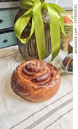 Freshly baked cinnamon roll placed on a linen tablecloth beside a glass jar tied with a green ribbon and a ceramic mug in a cozy kitchen setting Freshly baked cinnamon roll placed on a linen tablecloth beside a glass jar tied with a green ribbon and a ceramic mug in a cozy kitchen setting 137688041