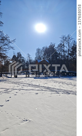 Snow-covered landscape with a wooden cabin on stilts surrounded by trees under a bright sun in a clear blue sky during winter season 137688058