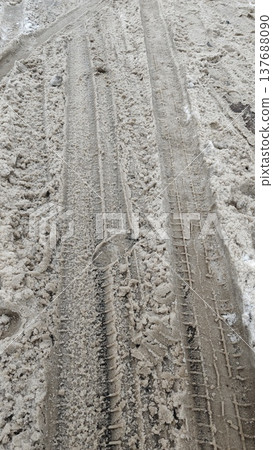 Tire tracks and footprints are visible on a snow-covered dirt road, indicating recent vehicle and pedestrian movement in a winter setting Tire tracks and footprints are visible on a snow-covered dirt road, indicating recent vehicle and pedestrian movement in a winter setting 137688090