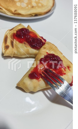 Pancakes filled with red berry jam on a white plate, fork inserted into one pancake, additional pancakes visible in the background on a separate plate Pancakes filled with red berry jam on a white plate, fork inserted into one pancake, additional pancakes visible in the background on a separate plate 137688091