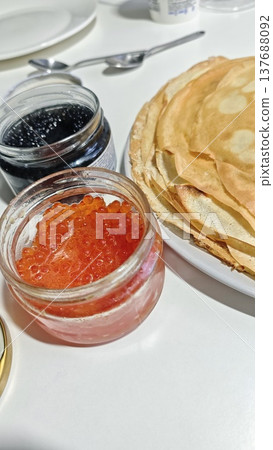 Close-up of traditional pancakes served with jars of red caviar and black caviar on a white table, showcasing a delicious culinary presentation Close-up of traditional pancakes served with jars of red caviar and black caviar on a white table, showcasing a delicious culinary presentation 137688092