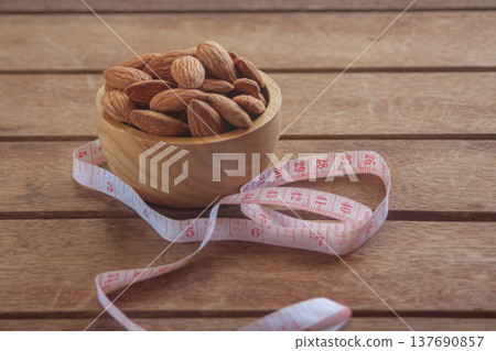 Almonds in a wooden cup on a wood plate with a measuring tape, symbolizing healthy diet control. Almonds in a wooden cup on a wood plate with a measuring tape, symbolizing healthy diet control. 137690857