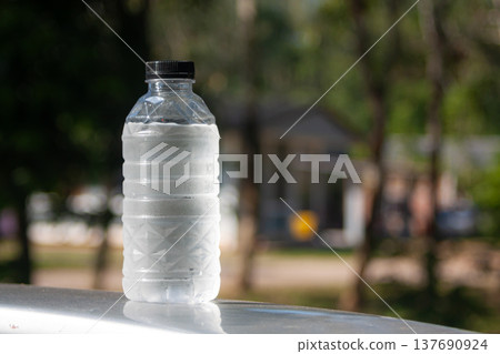 A cold plastic water bottle with condensation stands outdoors, catching sunlight against green blur. A cold plastic water bottle with condensation stands outdoors, catching sunlight against green blur. 137690924