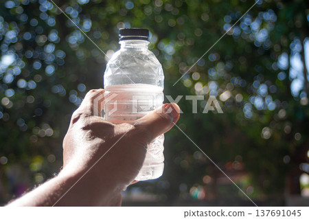 A hand holds a cold plastic water bottle outdoors, glowing in sunlight with soft green bokeh lit sun A hand holds a cold plastic water bottle outdoors, glowing in sunlight with soft green bokeh lit sun 137691045