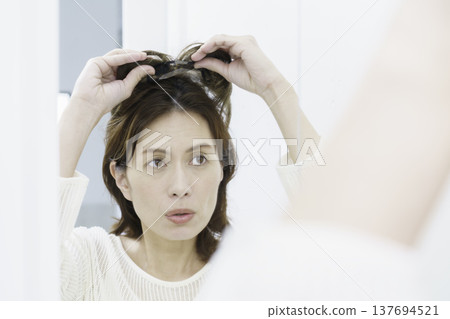 A middle-aged Japanese woman tries to put on a partial wig while looking in a mirror. A middle-aged Japanese woman tries to put on a partial wig while looking in a mirror. 137694521
