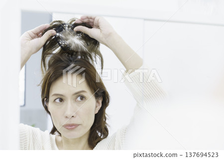 A middle-aged Japanese woman tries to put on a partial wig while looking in a mirror. A middle-aged Japanese woman tries to put on a partial wig while looking in a mirror. 137694523