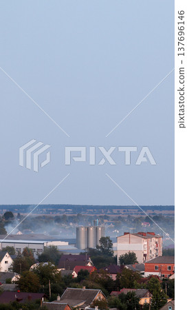 Vertical panorama of a small town with an industrial grain elevator and residential houses. View of a suburban landscape with a factory, apartment buildings, and private homes under evening sky 137696146