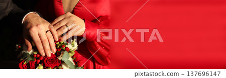 Close-up of a bride and groom's hands with wedding rings on a red rose bouquet. Wedding couple in red dress and dark suit, celebrating marriage. Large red copy space background Close-up of a bride and groom's hands with wedding rings on a red rose bouquet. Wedding couple in red dress and dark suit, celebrating marriage. Large red copy space background 137696147
