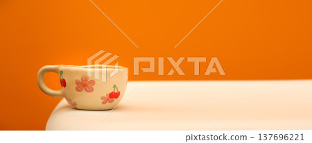 Minimalist still life with a ceramic cup on a white table. Beige handmade mug decorated with pink flowers and cherries against a solid orange background. Cozy morning and copy space concept 137696221