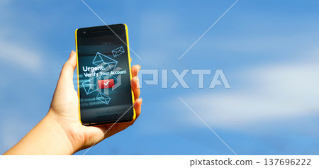 Cybersecurity and phishing alert concept. Hand holding a smartphone with an urgent account verification message and envelope icons on the screen against a blue sky background with copy space Cybersecurity and phishing alert concept. Hand holding a smartphone with an urgent account verification message and envelope icons on the screen against a blue sky background with copy space 137696222