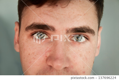 Close-up portrait of a young man with freckles and grey eyes looking at camera. Macro shot of male face with natural skin texture, bushy eyebrows, and light skin. Skin care and ophthalmology concept Close-up portrait of a young man with freckles and grey eyes looking at camera. Macro shot of male face with natural skin texture, bushy eyebrows, and light skin. Skin care and ophthalmology concept 137696224