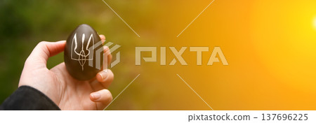 Patriotic Ukrainian Easter concept. A hand holding a dark Easter egg with a hand-drawn white Trident, Tryzub, the national symbol of Ukraine. background with bright sun flare and copy space 137696225