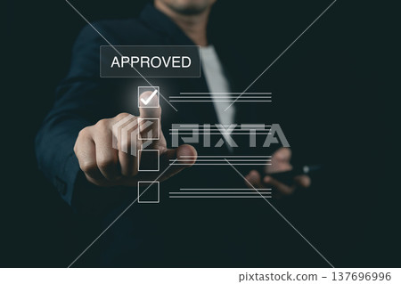 Businessman pointing finger at virtual checklist with approved sign on digital interface over dark background Businessman pointing finger at virtual checklist with approved sign on digital interface over dark background 137696996
