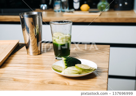 Mixing glass containing green liquid and cucumber slices sits on wooden counter next to metal shaker and lime. green juice cocktails, chlorophyll drinks, liquid wellness, vegetable-forward mixology Mixing glass containing green liquid and cucumber slices sits on wooden counter next to metal shaker and lime. green juice cocktails, chlorophyll drinks, liquid wellness, vegetable-forward mixology 137696999