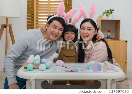 father, mother and daughter wearing bunny ears smiling happily while celebrating together at home on easter day. happy family holiday activity concept 137697083