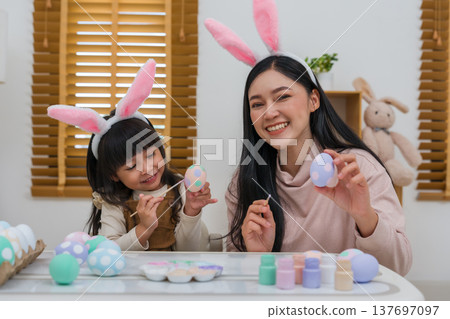 mother and preschool child girl wearing bunny ears painting and decorating egg together at home on easter day. happy family holiday activity concept mother and preschool child girl wearing bunny ears painting and decorating egg together at home on easter day. happy family holiday activity concept 137697097