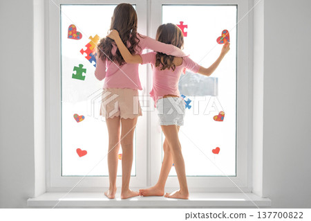Two girls standing by a bright window decorating glass with colorful puzzle pieces and hearts. Autism awareness, friendship, childhood support, inclusion and neurodiversity concept. 137700822