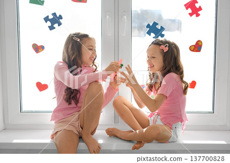 Happy children are sitting on the window sill, decorated with colorful pieces of puzzles and hearts. Girls give each other postcards on Autism Day Happy children are sitting on the window sill, decorated with colorful pieces of puzzles and hearts. Girls give each other postcards on Autism Day 137700828