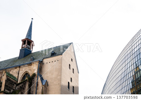 Antoniterkirche stands beside a modern urban building in Cologne, Germany. The church features a tall spire and traditional architecture, contrasting with the glass structure. Antoniterkirche stands beside a modern urban building in Cologne, Germany. The church features a tall spire and traditional architecture, contrasting with the glass structure. 137703598
