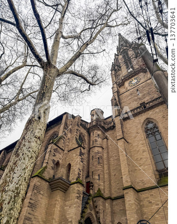 Herz Jesu Church in Cologne, Germany. The image captures the church's tall spire and intricate stonework, surrounded by bare trees against a cloudy sky. Herz Jesu Church in Cologne, Germany. The image captures the church's tall spire and intricate stonework, surrounded by bare trees against a cloudy sky. 137703641