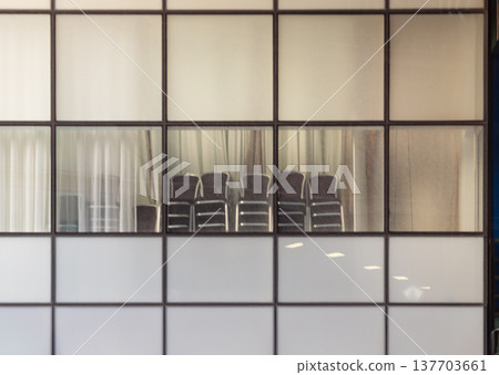 A study in quiet stillness, this tableau of functional simplicity is framed by a grid of glass and metal. Frosted lower panels obscure the floor, while clear panes above reveal utilitarian chairs 137703661