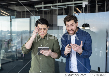 Two diverse male colleagues discussing business issues and expressing frustration over work challenges while looking at a digital tablet in a modern office environment Two diverse male colleagues discussing business issues and expressing frustration over work challenges while looking at a digital tablet in a modern office environment 137703685