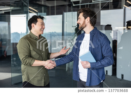 Two male businesspeople from different ethnicities happily shaking hands, building trust and cooperation during a successful negotiation in a modern glass office Two male businesspeople from different ethnicities happily shaking hands, building trust and cooperation during a successful negotiation in a modern glass office 137703688