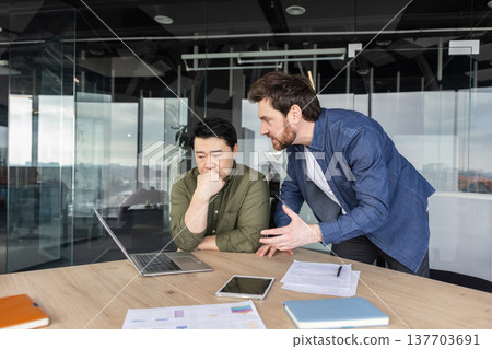 Two diverse male employees discussing a project, one appearing stressed and overwhelmed while the other expresses frustration and disagreement in a modern office meeting room setting 137703691