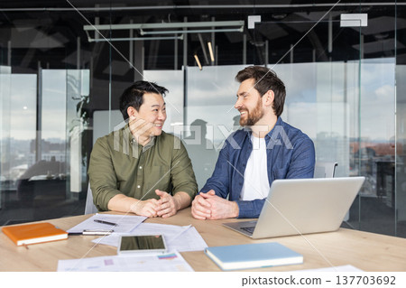 Two diverse male business partners discussing work and smiling while sitting at a table with papers and a laptop in a contemporary meeting room, showing teamwork and partnership Two diverse male business partners discussing work and smiling while sitting at a table with papers and a laptop in a contemporary meeting room, showing teamwork and partnership 137703692