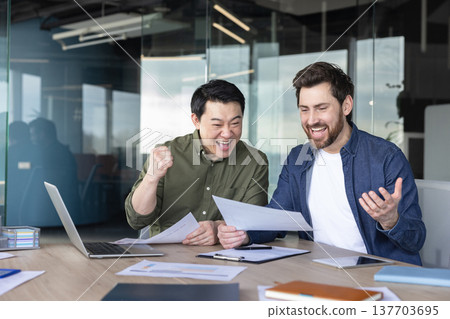 Two excited diverse businessmen celebrating a successful deal or project outcome, holding documents and expressing joy while working together at a meeting table in a contemporary office 137703695