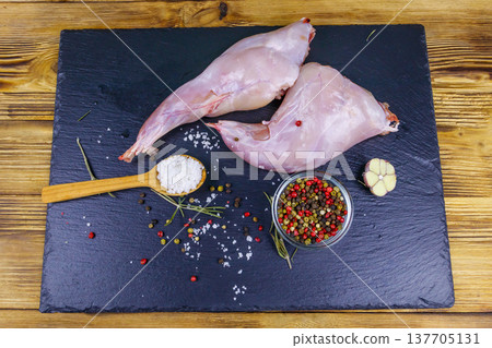 Raw rabbit legs with spices on a black slate cutting board on wooden table. Top view Raw rabbit legs with spices on a black slate cutting board on wooden table. Top view 137705131