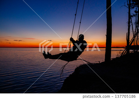 Silhouette of a man on rope swing above river at sunset Silhouette of a man on rope swing above river at sunset 137705186